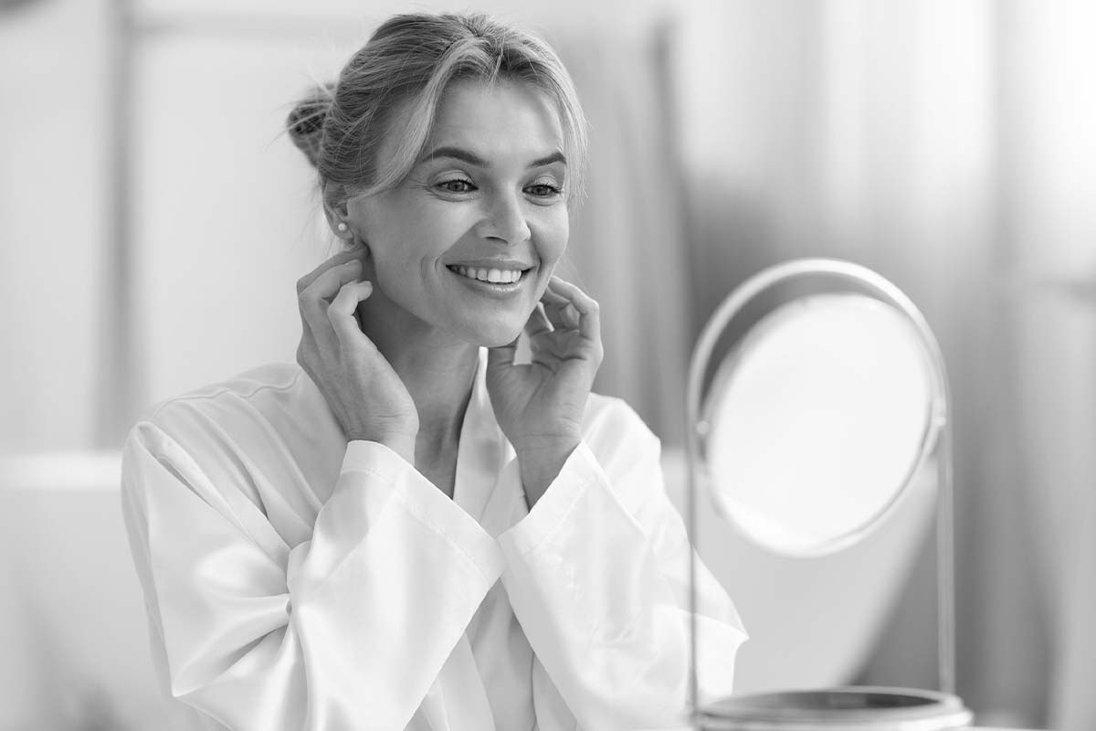 a woman sitting in a robe looking at her reflection in a mirror and smiling after eyelid surgery in Dallas
