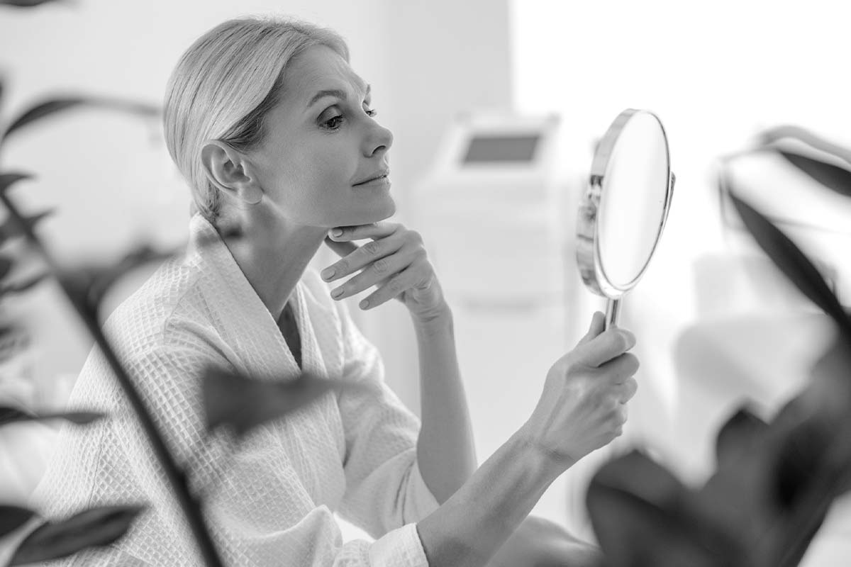 an older woman sitting in a robe and looking at herself in a hand mirror after getting a neck lift in Dallas